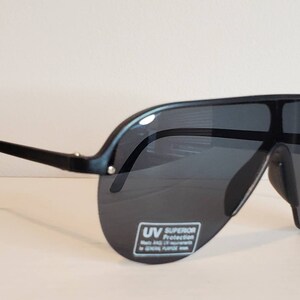 Op de afbeelding: Zwarte zonnebril met een wrap-around ontwerp. De zonnebril heeft donkere glazen en een dik, gebogen montuur. De glazen hebben een sticker met de tekst "UV Superior Protection". Ontworpen voor algemeen gebruik.