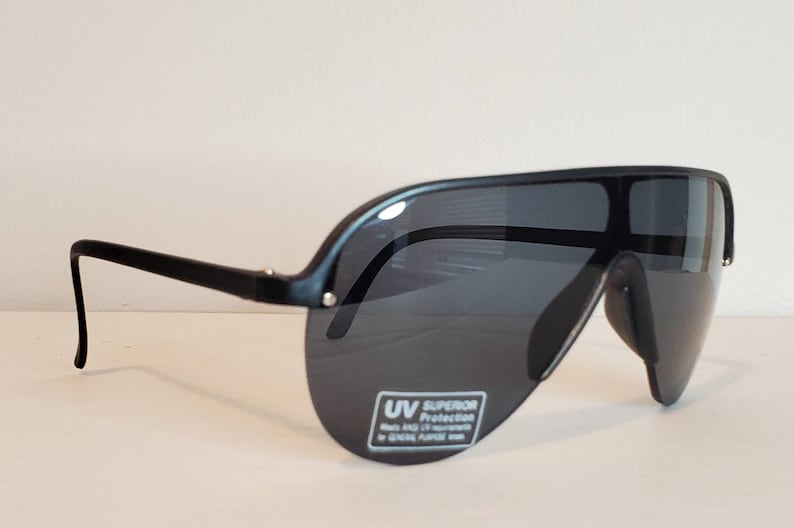 Op de afbeelding: Zwarte zonnebril met een wrap-around ontwerp. De zonnebril heeft donkere glazen en een dik, gebogen montuur. De glazen hebben een sticker met de tekst "UV Superior Protection". Ontworpen voor algemeen gebruik.
