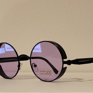May include: Black round sunglasses with a thick black frame and purple lenses. The sunglasses have a metal bridge and a UV 400 protection label on the right lens.