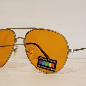 May include: Aviator-style sunglasses with amber-colored lenses and a gold-tone metal frame. The lenses have a UV Amber Protection label and block blue light. The temples are silver with black ear pieces.