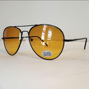 May include: Aviator sunglasses featuring a thin black metal frame and amber-tinted lenses. A small white sticker on the right lens displays the text PC Lens, UV Protection, and Spring Temple. The design includes a double bridge and nose pads.
