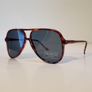 May include: Tortoiseshell-framed sunglasses with dark lenses. The aviator-style sunglasses have a double bridge and a sticker that reads "POLARIZED LENSES". The frames are brown with a mottled pattern.