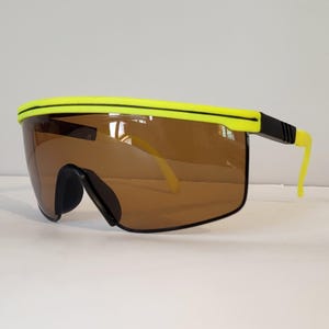 Puede incluir: Gafas de sol de estilo retro con montura amarillo neón y lentes marrones. Las gafas tienen un diseño envolvente con detalles negros en las patillas y el puente nasal. Las patillas también son de color amarillo neón.