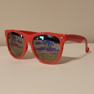 Puede incluir: Gafas de sol rojas con lentes espejadas que reflejan una escena de playa tropical. Las monturas son de un rojo brillante, y las lentes muestran palmeras, arena y un cielo colorido. Un accesorio elegante.