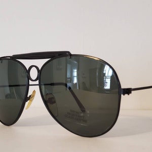 May include: Black aviator sunglasses with a dark green lens and a metal frame. The sunglasses have a double bridge and a metal bar across the top of the frame.