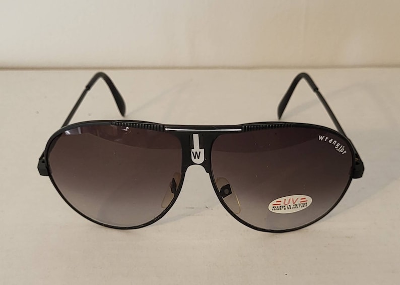 May include: Black Wrangler aviator sunglasses with gradient lenses. The sunglasses have a black frame with the Wrangler logo on the right lens and a white "W" on the bridge. A small UV protection sticker is on the right lens.