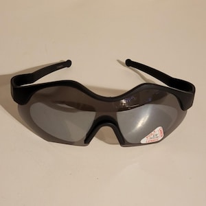 Puede incluir: Gafas de sol deportivas negras con lentes espejadas. Las gafas tienen un diseño envolvente con montura y patillas negras. Una pequeña pegatina en la lente dice "POLY UV PROTECTIVE LENSES".