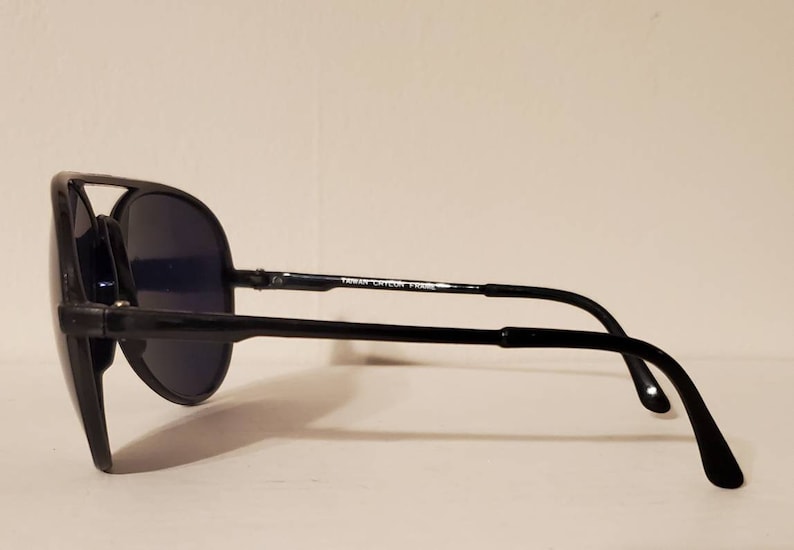 May include: Black aviator sunglasses with a black frame and dark lenses. The sunglasses have a metal bar on the bridge of the nose and the text "Taiwan Cycon Frame" is printed on the bar.