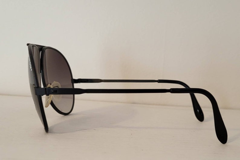 May include: Black aviator sunglasses with gradient lenses. The sunglasses feature a sleek, minimalist design with a black frame and temples. The lenses transition from dark to light, offering a stylish and functional accessory.
