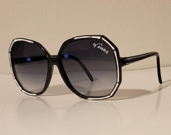 Puede incluir: Gafas de sol negras con montura geométrica y lentes degradadas. Las gafas tienen detalles blancos y la palabra "Venus" en escritura cursiva en la lente superior derecha. Las patillas son finas y negras.