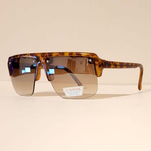 May include: Brown and tan sunglasses with a tortoise shell pattern on the frames. The sunglasses have a rectangular shape with a flat top and a curved bottom. The lenses are a gradient brown color. A small sticker on the lens reads "UV PROTECTION".