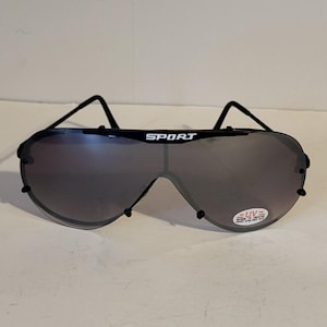 May include: Black shield-style sunglasses featuring a single continuous dark lens with the word SPORT printed in white on the top center. A small oval sticker on the lower right lens reads UV with additional text below. The frame has thin black arms.