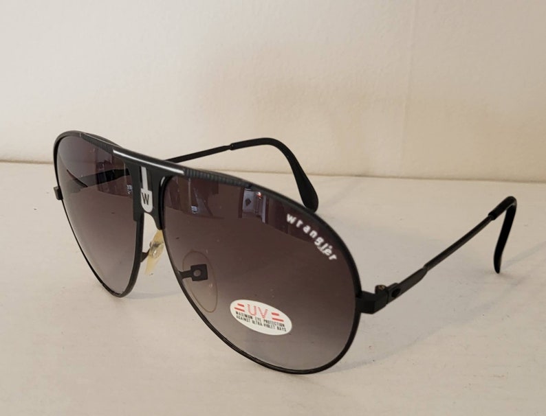 May include: Black Wrangler aviator sunglasses with gradient lenses. The sunglasses have a black frame with the Wrangler logo on the side. A UV protection sticker is visible on the lens.