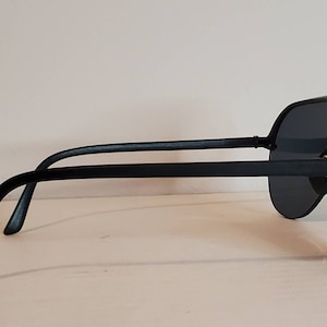 Op de afbeelding: Zwarte zonnebril met donkere glazen. De zonnebril heeft een dik, rechthoekig montuur en rechte pootjes. De glazen zijn donkergrijs en het montuur is glanzend zwart. De zonnebril wordt van opzij bekeken.