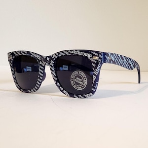 May include: Dark navy sunglasses with black lenses. The frames are covered in a white text pattern. The lenses have a circular sticker that reads "SUPER DARK LENS IMPACT RESISTANT NO GLARE".