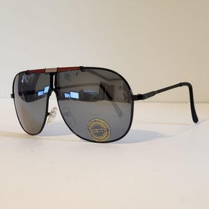 Op de afbeelding: Zwarte pilotenzonnebril met zilverkleurige spiegelglazen. De zonnebril heeft een bruin accent op de bovenste stang en een gouden sticker op de lens. De monturen zijn zwart en de pootjes zijn dun.
