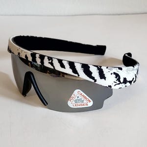May include: Sport sunglasses with a black and white zebra print headband and gray lenses. The lenses have a sticker that reads "Poly Carbon Lenses UV Protection". The sunglasses have a black frame.