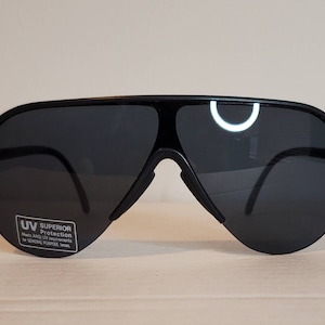 Op de afbeelding: Zwarte piloten zonnebril met donkere glazen. De zonnebril heeft een dik zwart montuur en een brug over de neus. Een kleine sticker op de lens luidt "UV Superior Protection".