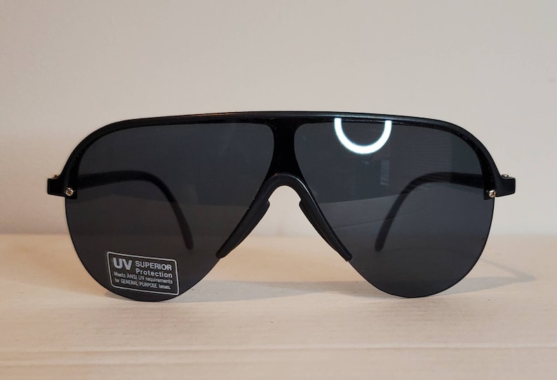 Op de afbeelding: Zwarte piloten zonnebril met donkere glazen. De zonnebril heeft een dik zwart montuur en een brug over de neus. Een kleine sticker op de lens luidt "UV Superior Protection".
