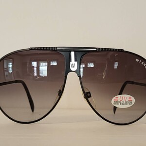 May include: Black Wrangler aviator sunglasses with gradient lenses. The sunglasses have a black frame with a white stripe and the Wrangler logo on the side. A sticker indicates maximum UV protection.