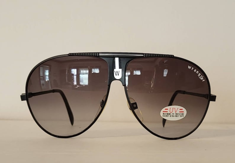 May include: Black Wrangler aviator sunglasses with gradient lenses. The sunglasses have a black frame with a white stripe and the Wrangler logo on the side. A sticker indicates maximum UV protection.