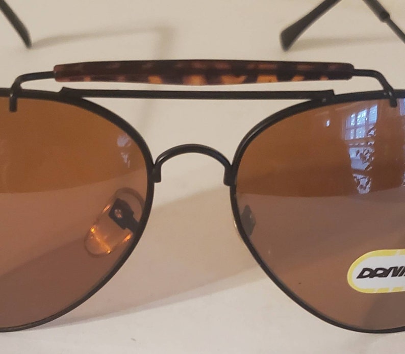 May include: Black aviator sunglasses with brown lenses and a tortoise shell brow bar. The sunglasses have a metal frame and a 'DONNA' label on the right lens.