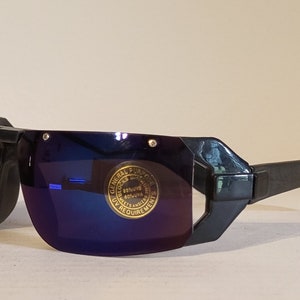 May include: Black sunglasses with a wraparound design and blue mirrored lenses. The sunglasses have a unique, angular frame and a small sticker on the lens. The lenses block 80% UVA/UVB rays.