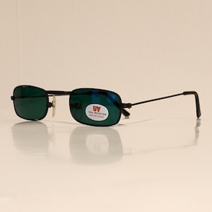 May include: Rectangular sunglasses with a black metal frame and green lenses. The lenses have a small white sticker that reads "UV 100% PROTECTION". The sunglasses are displayed on a white surface.