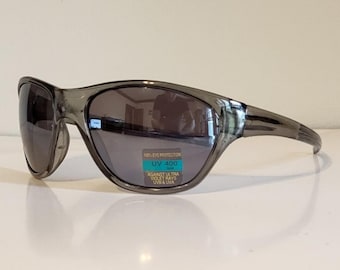 Może przedstawiać: Szare sportowe okulary przeciwsłoneczne z ciemnymi soczewkami. Okulary przeciwsłoneczne mają owijany design i zakrzywioną oprawkę. Mała naklejka na soczewce głosi: "100% EYE PROTECTION UV 400 AGAINST ULTRA VIOLET RAYS UVA & UVB".