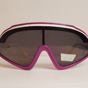 Può includere: Occhiali da sci viola e bianchi con lente in policarbonato. Gli occhiali hanno un design futuristico e sono in plastica.