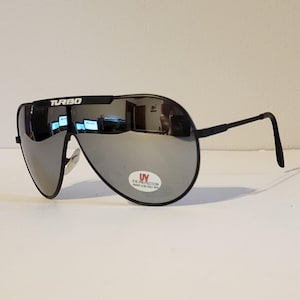 Może przedstawiać: Czarne okulary przeciwsłoneczne typu aviator z lustrzanymi soczewkami. Okulary mają czarną oprawkę, a na górze oprawki nadrukowano napis "TURBO". Mała naklejka na soczewce wskazuje na ochronę oczu przed promieniowaniem UV.