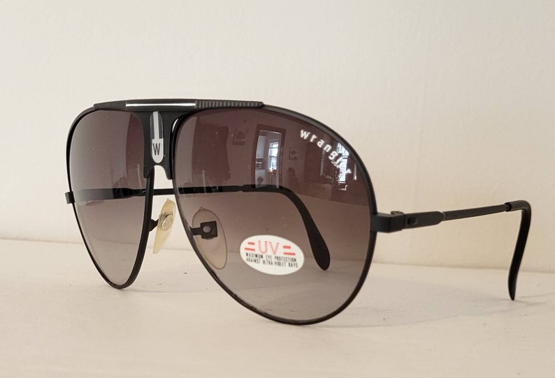 May include: Black Wrangler aviator sunglasses with gradient lenses. The sunglasses have a black frame with the Wrangler logo on the lens. A sticker on the lens reads "UV Maximum Eye Protection".
