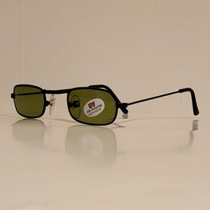May include: Black rectangular sunglasses with green lenses. The glasses have a thin black frame and a small sticker on the right lens that reads "UV 100% PROTECTION". The sunglasses are displayed on a white surface.