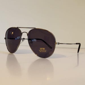 May include: Aviator-style sunglasses with dark lenses and a thin, metal frame. The lenses have a slight gradient and the frame features a double bridge. The sunglasses have the text "UV 100 EYE PROTECTION" printed on the lens.