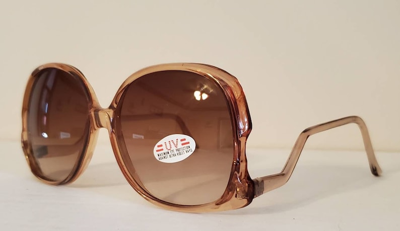 May include: Oversized, light brown sunglasses with brown tinted lenses. The sunglasses have a rounded shape and a sticker that reads "UV Maximum Eye Protection Against Ultra Violet Rays". The arms are a light brown color.