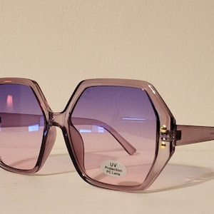 May include: Oversized, hexagonal sunglasses with a gradient purple lens. The frames are a translucent lavender color, with gold-colored accents on the hinges. The sunglasses have UV protection and PC lenses.
