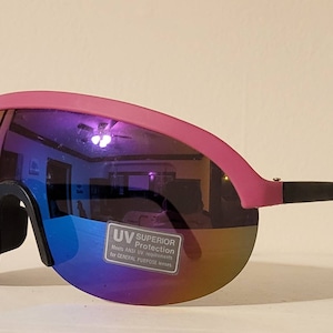 Puede incluir: Gafas de sol rosas y negras con una lente grande y curvada. Las lentes tienen un acabado espejado y multicolor. La montura es rosa con patillas negras. Una pegatina en la lente dice "UV Superior Protection".