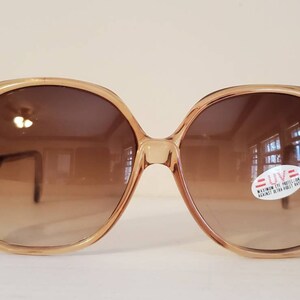 May include: Oversized, vintage sunglasses with a light brown frame and gradient brown lenses. A sticker on the lens reads "UV: MAXIMUM EYE PROTECTION AGAINST ULTRA VIOLET RAYS". The sunglasses have a retro, 70s style.