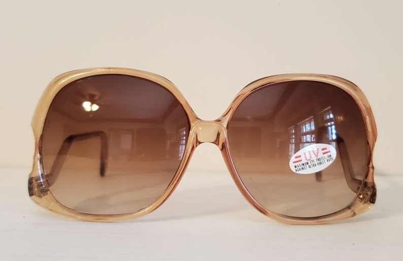 May include: Oversized, vintage sunglasses with a light brown frame and gradient brown lenses. A sticker on the lens reads "UV: MAXIMUM EYE PROTECTION AGAINST ULTRA VIOLET RAYS". The sunglasses have a retro, 70s style.