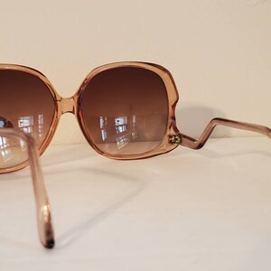 May include: Oversized, light brown sunglasses with a unique zig-zag temple design. The lenses are a gradient brown, and the frames have a rounded, retro shape. The sunglasses are set against a neutral background.