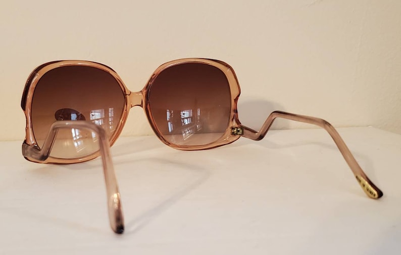 May include: Oversized, light brown sunglasses with a unique zig-zag temple design. The lenses are a gradient brown, and the frames have a rounded, retro shape. The sunglasses are set against a neutral background.
