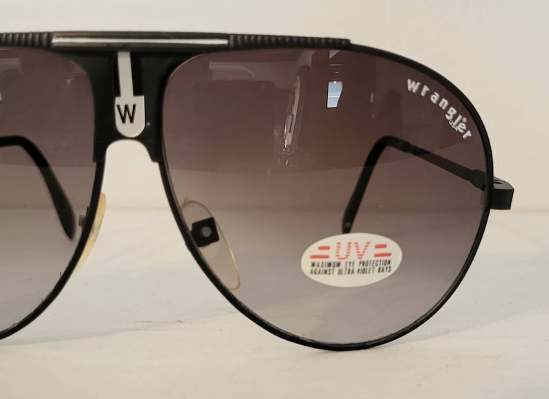 May include: Black Wrangler sunglasses with gradient lenses. The sunglasses have a black frame with a white stripe and the letter "W" on the bridge. The lenses have a UV protection sticker.