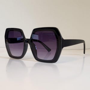 May include: Black oversized sunglasses with a geometric, octagonal frame. The lenses have a gradient tint, transitioning from dark at the top to lighter at the bottom. The sunglasses are set against a white background.