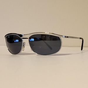 May include: Silver-framed sunglasses with dark lenses. The sunglasses have a sleek, modern design with a curved top bar and straight temples. The lenses are oval-shaped and reflect the surrounding environment.