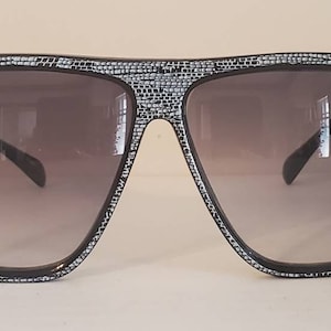 Puede incluir: Gafas de sol negras con un marco estampado en blanco. Las lentes son de color gris oscuro.