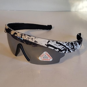 May include: Sport sunglasses with a white and black patterned frame and dark gray lenses. The sunglasses have a black nose bridge and a black headband. A sticker on the lens reads "Poly Carbon Lenses".