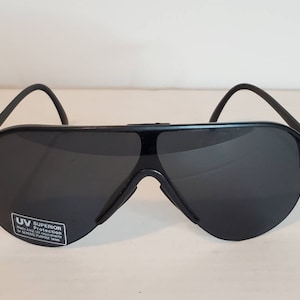 Op de afbeelding: Zwarte zonnebril met een gebogen bovenframe en donkere glazen. De zonnebril heeft een afgeronde vorm en een kleine UV-beschermingssticker op de linker onderlens. De pootjes zijn dun en gebogen.