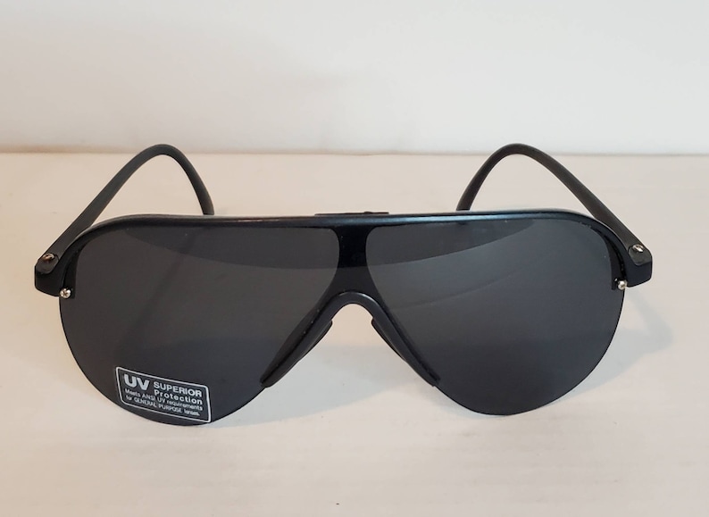 Op de afbeelding: Zwarte zonnebril met een gebogen bovenframe en donkere glazen. De zonnebril heeft een afgeronde vorm en een kleine UV-beschermingssticker op de linker onderlens. De pootjes zijn dun en gebogen.