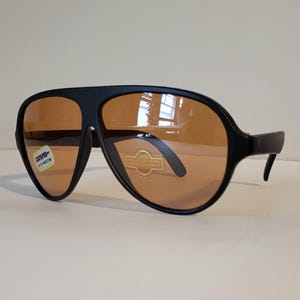 May include: Black aviator sunglasses with orange-tinted lenses. The sunglasses have a thick, matte black frame and a classic teardrop shape. The lenses have a slight gradient. The sunglasses have a sticker that says "UV400 Eye Protection".
