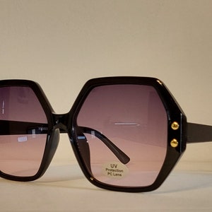 May include: Black oversized sunglasses with a hexagonal frame and gradient pink lenses. The sunglasses have gold accents on the frame and a label that reads "UV Protection PC Lens".